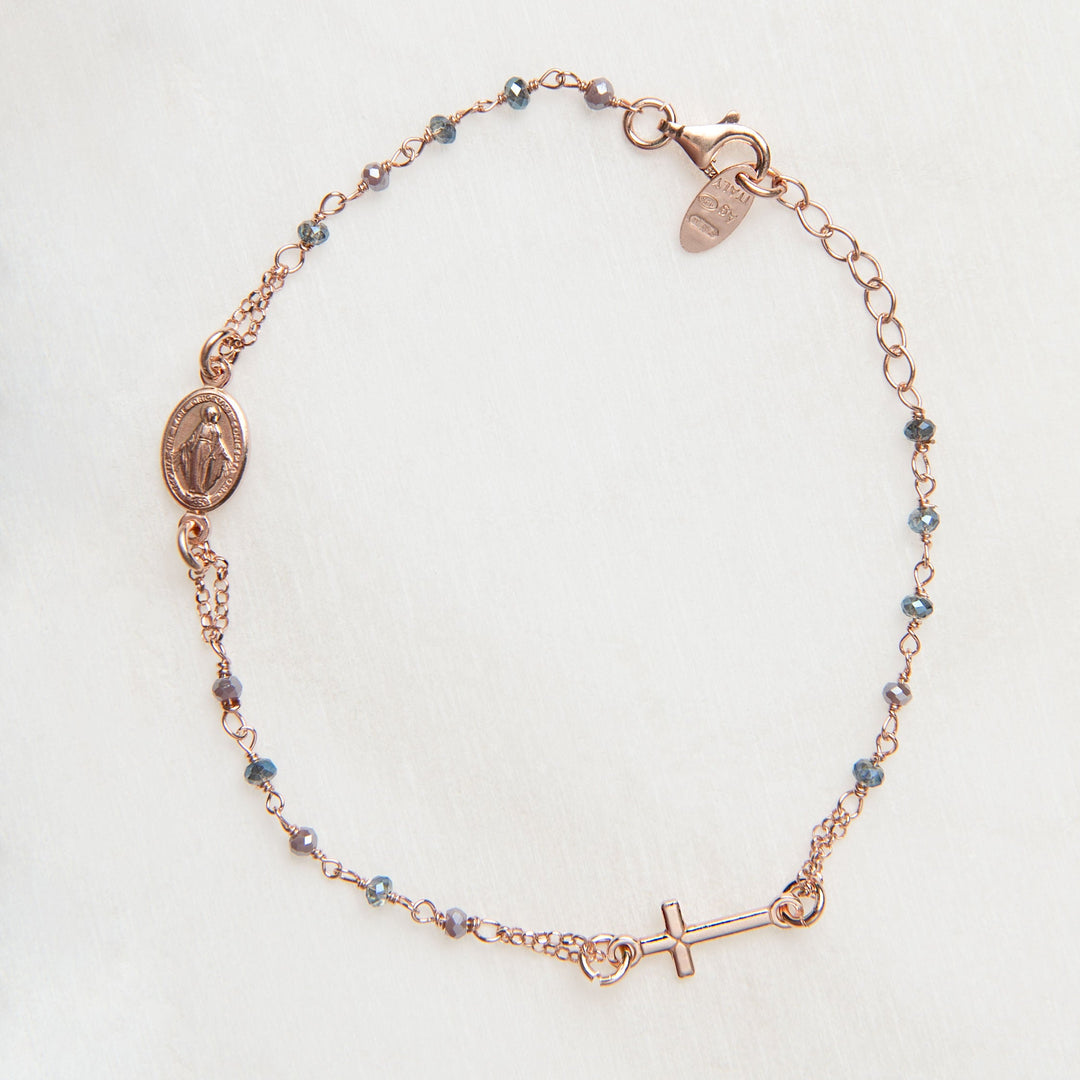 A delicate rose gold chain bracelet featuring a Miraculous Medal charm, a small cross charm, and tiny blue beads spaced along the chain, laid flat on a light textured background.