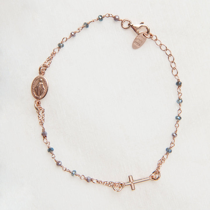 A delicate rose gold chain bracelet featuring a Miraculous Medal charm, a small cross charm, and tiny blue beads spaced along the chain, laid flat on a light textured background.